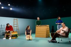 Ensemblen i föreställningen "Bön för idioter" poserar på scenen med rekvisita, inklusive en stege och en stor hamburgermodell.