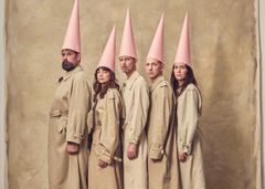 Bild av fem personer i beige rockar och rosa strutformade hattar mot en brun bakgrund.