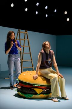 Två personer på en scen, en står bredvid en stege, den andra sitter på en stor hamburgerekvisita.