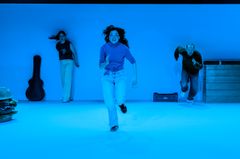 Bild på tre personer som springer entusiastiskt på en blåtonad scen. En gitarr och en stor hamburgare syns i bakgrunden.