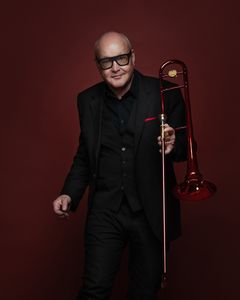 En man i svart kostym håller en röd trombon mot en mörkröd bakgrund.