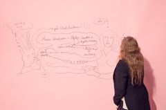 Kvinna i mörk kavaj ritar på rosa vägg.