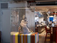 Keramisk skulptur i glasmonter på randigt podium, stående i en modern, luftigt upplyst lokal.