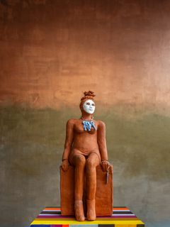 En skulptur av en figur sittande på en färgglad piedestal, mot en tvåtonad bakgrund.