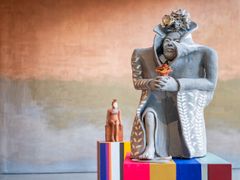 Skulptur av en person som sitter och håller en blomma, omgiven av färgglada piedestaler.