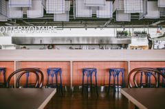 Interiör av restaurang Dotter Voltaire med moderna detaljer, inklusive blå barstolar och ett öppet kök.