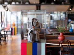Keramisk skulptur föreställande en person med en stiliserad, rustik design. Figuren omfamnar en liten blomma och är placerad på en färgglad piedestal i en utställningsmiljö.