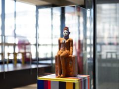En keramisk skulptur i en glasmonter, placerad på en flerfärgad piedestal. Skulpturen föreställer en sittande figur.