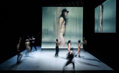 Ensemblen uppträder på scenen framför en stor bildskärm med konstnärliga visualer.