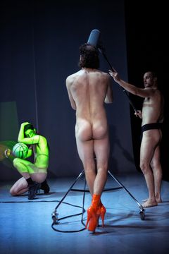 Tre personer på en scen, en med en boll, en med ett stativ och en med orangea klackar.