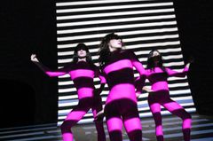 Tre personer i färgglada kostymer står framför en bakgrund med horisontella ränder.