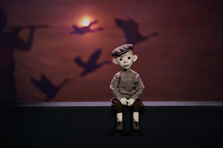 En marionettdocka föreställande Nils Holgersson sitter på en kant, med silhuetter av flygande gäss i bakgrunden.