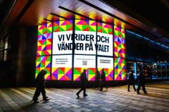 Skylt med texten "Vi vrider och vänder på valet" på en byggnad med färgglad geometrisk bakgrund. Flera personer går förbi på trottoaren.