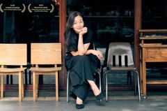 Lee Chia-Ying sitter leende på en stol framför en butik med utmärkelser på fönstret.