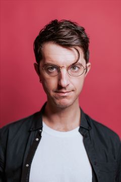 Vincenzo Latronico med glasögon och mörkt hår, bär en vit t-shirt och svart skjorta, framför en röd bakgrund.