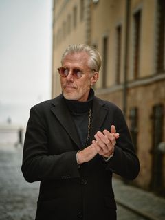 Carl-Johan Vallgren i mörk rock och solglasögon, stående utomhus framför en byggnad.