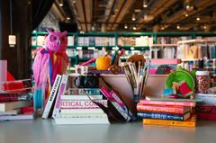 Böcker, målarsaker och en rosa uggla på ett skrivbord i ett bibliotek.