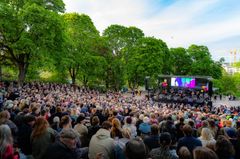 En stor publik sitter framför en utomhusscen i en grönskande park under en föreställning.