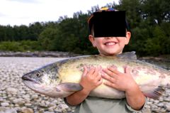 Ett barn står vid en stenig flodstrand och håller en stor fisk med båda händerna. Barnets ansikte är täckt.