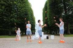Fyra personer jonglerar med orange bollar i en park, omgivna av träd och vita kuber.