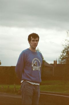 En man står utomhus med molnig himmel i bakgrunden, klädd i en blå sweatshirt och jeans.