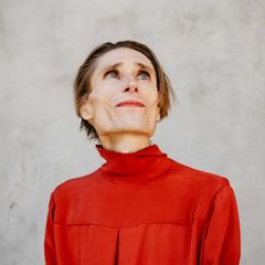 Porträtt av Madame Nielsen i röd polotröja med blicken riktad uppåt, mot en ljusgrå bakgrund.