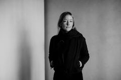 Svartvitt porträtt av Eva Åsefeldt i mörk jacka och halsduk, stående med händerna i fickorna mot en ljus vägg.