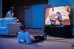 Repetitionsbild för "Sisyfos i vardagsrummet" med tre personer i en teaterscen där en står, en sitter på golvet och en visas på TV-skärmen.