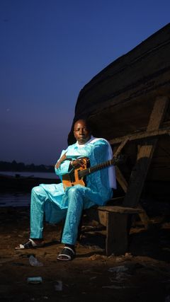 Vieux Farka Touré sitter med gitarr utanför en båt i skymningen.