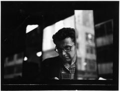 Svartvitt fotografi av Saul Leiter med glasögon, fotograferad genom en glasruta med mörka partier och reflektioner runt ansiktet.