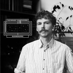 Svartvitt porträtt av Aron Landahl med mustasch och randig skjorta framför bokhyllor. Uppe till vänster syns logotypen för Kulturhuset Stadsteaterns Bilderbokspris.