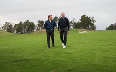 Två män promenerar på en golfbana med träd i bakgrunden.