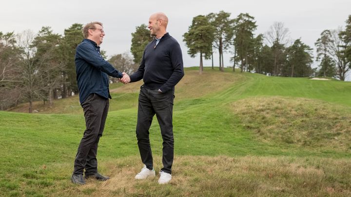 Två medelålders män står på en golfbana och ler och skakar hand.