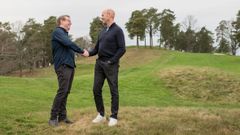 Två män står på en gräsklädd kulle på en golfbana och skakar hand, omgivna av träd i bakgrunden.