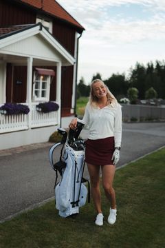 Kvinna klädd i golfkläder står bredvid en golfvagn på en gräsmatta framför ett hus.