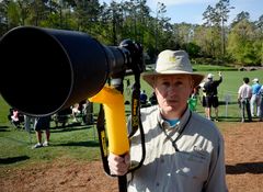 En manlig fotograf i beige hatt och ett stort kameraobjektiv riktat in mot kameran, står på golfbanan Augusta National under The Masters.