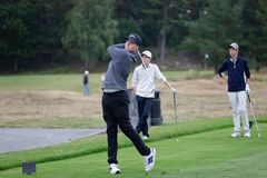 En ung golfspelare svingar en klubba på tee på en golfbana, medan två andra unga spelare tittar på i bakgrunden.