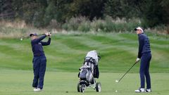 Två unga golfare spelar på en grön golfbana; en slår ett slag medan den andra står bredvid en golfvagn.