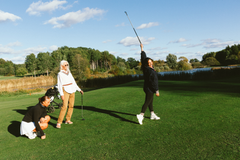 Tre kvinnor står på en golfbana; en kvinna svingar en golfklubba genom luften.