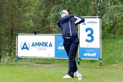 Ung golfspelare i blå kläder svingar en klubba, med skyltar för ANNIKA Invitational Europe och Ahlsell i bakgrunden på en golfbana.