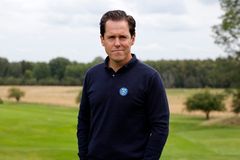 En man står på en golfbana med träd i bakgrunden, iklädd en mörk tröja med SGF-logotyp.