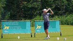 Ung golfspelare i grå tröja och mörkblå shorts slår ett utslag på en golfbana, med skyltar för "Svenska Juniortouren" i bakgrunden.