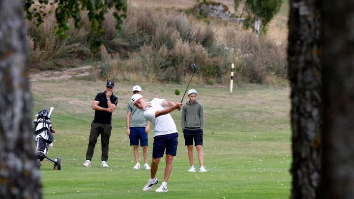 En pojke slår en golfboll med en driver medan tre andra personer tittar på, på en golfbana med gräs och buskar i bakgrunden.