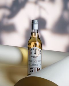 Flaska gin med etikett "Chestnut Aged" står på ett bord, omgiven av böjda papper.