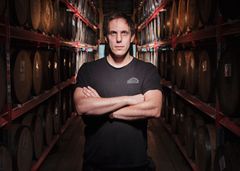 Oskar Bruno i svart t-shirt står med armarna i kors i en lagerlokal fylld med träfat.