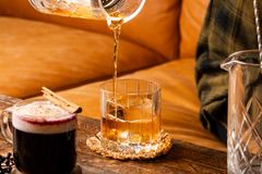 En klar dryck hälls i ett glas med isbitar på ett träbord, bredvid en mörk dryck med grädde och en kanelstång.