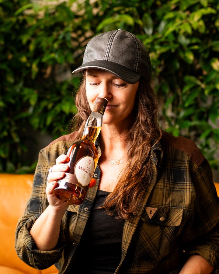 Kvinna%20i%20keps%20doftar%20p%E5%20en%20flaska%20calvados%2C%20framf%F6r%20gr%F6nskande%20bakgrund.