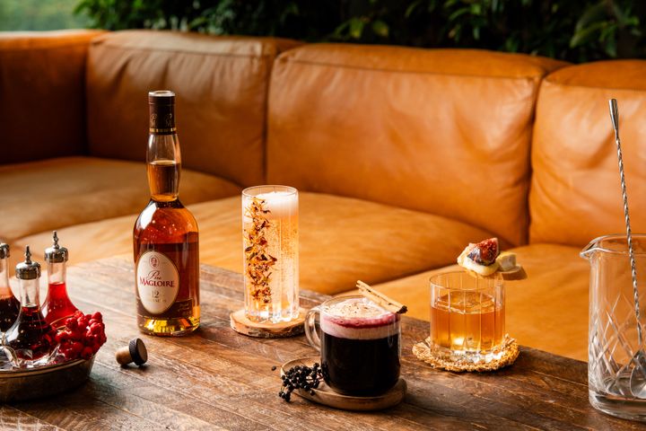 En flaska Père Magloire bredvid tre olika drinkar på ett träbord framför en brun soffa.