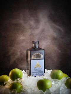 Flaska med Agitator Ice Cider Whisky omgiven av is och gröna äpplen.