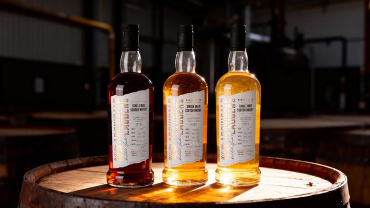 Tre flaskor med whisky står på en trätunna i ett svagt upplyst rum.